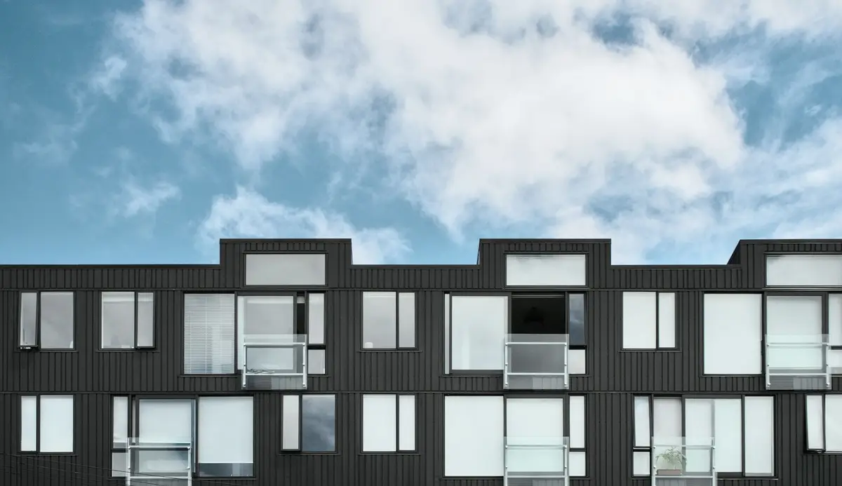 A dark modular residential facade with large windows beneath a wide sky with clouds